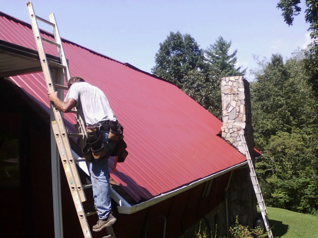 Roofer on a ladder beside a red metal roof and stone chimney — Soffit & Fascia Repair by Roofing Brothers in Dewey
